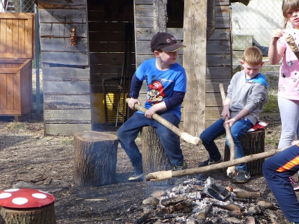 Kinder sitzen auf Baumstammabschnitten um ein Feuer herum und bräteln Stockbrote