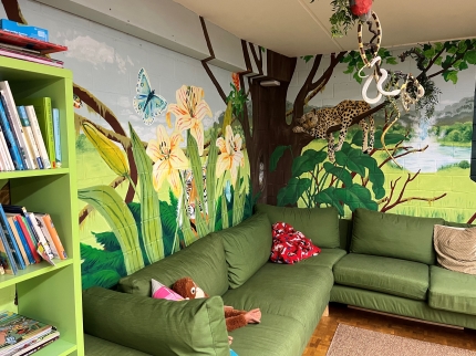 Grünes Sofa mit Dschungelwaldbild im Hintergrund und Büchergestell am linken Rand des Bildes