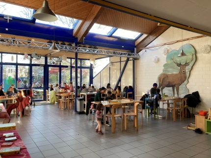 Grosser Raum mit sechser Tischen, an welchen Erwachsene und Kinder sitzen und Brettspiele spielen