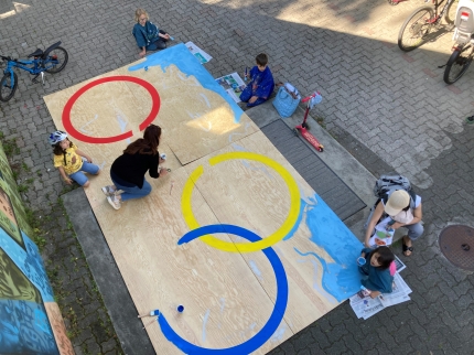 Verschiedene Kinder und Erwachsene bemalen eine riesengrosse Holzplatte mit den Olympischen Ringen