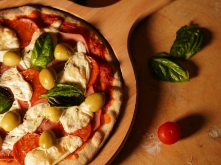 Ein Pizza auf einem runden Holzbrett mit Oliven, Basilikum und Tomaten.