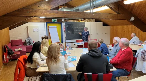 Eine Gruppe Erwachsener Sitzt um einen Tisch herum und hört der Deutschlehrerin zu